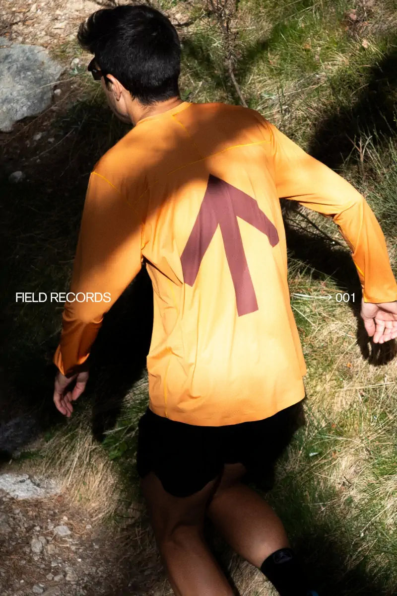 A runner wearing a bright orange long-sleeve running t-shirt, walking outdoors on grass and rocks in dappled sunlight.